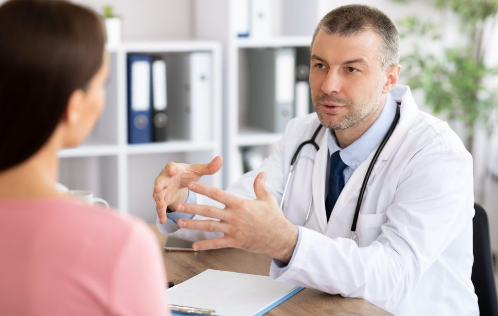 Middle aged experienced doctor talking to his patient Visiting Physician Concept. Portrait of experienced middle aged doctor talking to woman, explaining diagnosis and treatment. Therapist consulting young patient sitting at table in hospital